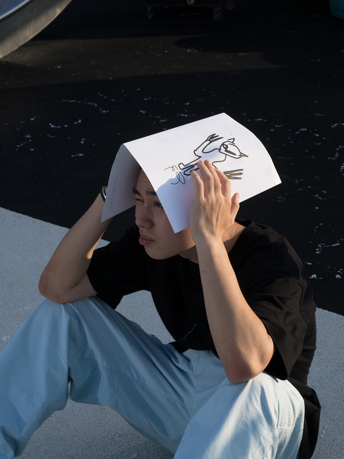 A person holds a folded paper with an illustration over his head while sitting on the ground. 